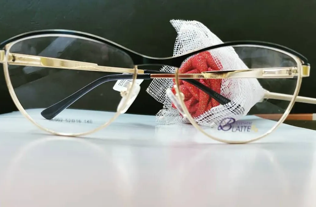 Black and gold cat-eye glasses on a white table in front of a deep green wall, with a red fabric rose behind